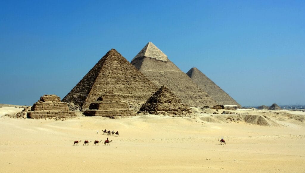 The iconic Pyramids of Giza under a clear blue sky with camels traversing the sandy desert.